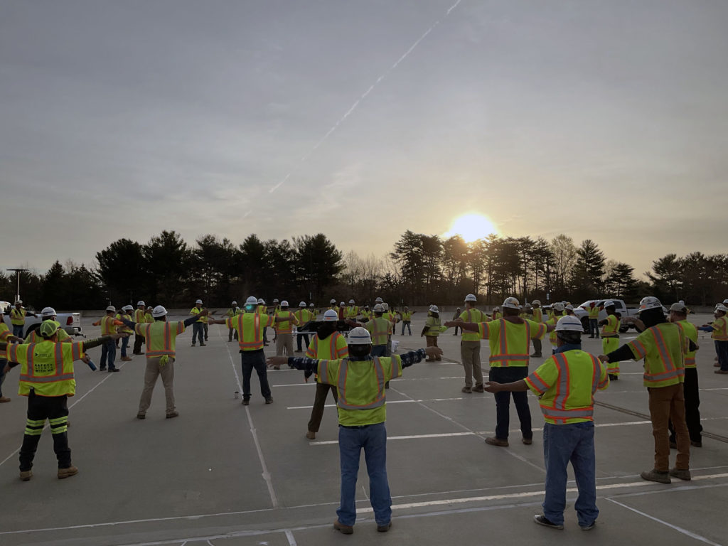 Workers standing apart stretching in a parking lot while the sun rises